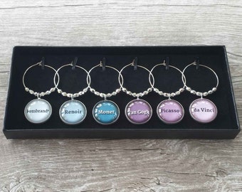 May include: A set of six wine charms with silver hoops and beaded accents. Each charm features a circular image with the names of famous artists: Rembrandt, Renoir, Monet, Van Gogh, Picasso, and da Vinci. The charms are displayed in a black box.