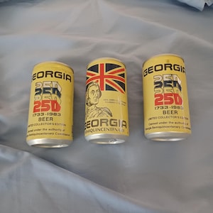 May include: Three yellow aluminum beer cans commemorating the Georgia Semiquincentenary 1733-1983. Two cans feature the text Georgia Beer 250 Limited Collector's Edition. The center can displays a Union Jack flag and a portrait of Gen. James Oglethorpe.
