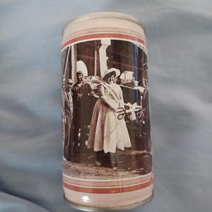 May include: A cylindrical metal beverage can featuring a sepia-toned photographic print of a group of people, including a central figure wearing a sash and hat, participating in a ribbon-cutting ceremony. The can has light pink top and bottom rims.