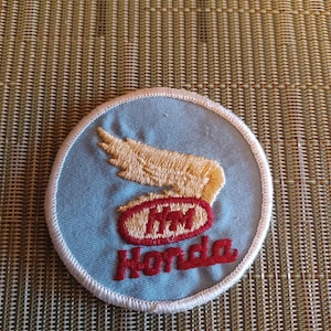 May include: A round, light blue Honda patch with a cream-colored wing design and the Honda logo in red. The patch has a white border and is likely for a jacket or bag.