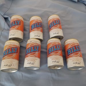 May include: Seven Billy Beer cans with orange and white labels. Each can features the blue Billy Beer logo, text stating the beer was brewed for Billy Carter, and a printed signature. The cans are arranged in a group on a light blue fabric surface.