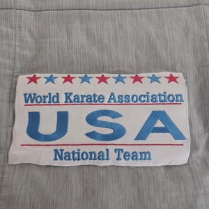 May include: A rectangular white fabric patch featuring embroidered text and graphics. At the top, a row of alternating red and blue stars. Below, the blue embroidered text reads World Karate Association, USA, and National Team, with red lines.