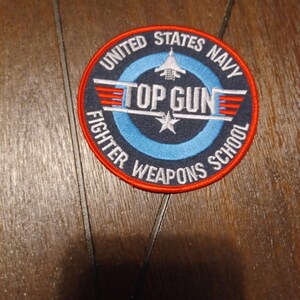 May include: Circular patch with the text "UNITED STATES NAVY TOP GUN FIGHTER WEAPONS SCHOOL". The patch has a red border, a blue inner circle, and white text and design elements, including an airplane and a star.