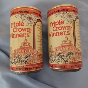 May include: Two cylindrical metal beverage cans featuring a cream and red design. The text reads Triple Crown Winners, Preakness, and May 11th thru 21st. The graphic depicts a horse racing scene with jockeys and a building, plus a list of horse names.