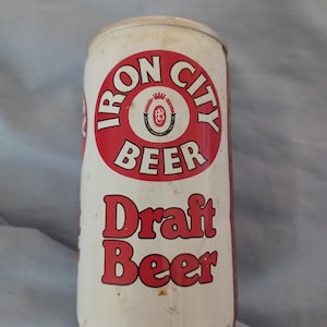May include: A white metal beer can featuring a red circular logo with the text Iron City Beer and a central crest. Below the logo, the words Draft Beer are printed in red. The can has a metallic top and bottom rim with visible signs of wear.