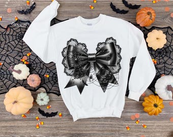 May include: White sweatshirt featuring a large black bow design with spiderwebs and lace detailing. The bow has a satin finish and is decorated with bats and spiders. The sweatshirt is part of a Halloween-themed collection.