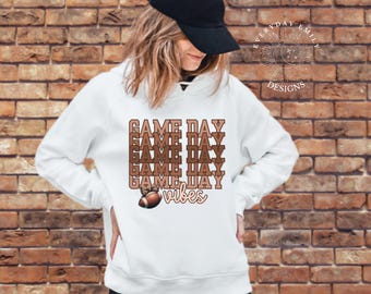 May include: White hoodie with the text "GAME DAY" in a stacked design, along with a football graphic and the word "vibes". The design is in shades of brown and tan. The hoodie is worn by a person wearing a black baseball cap.