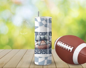 May include: A stainless steel tumbler with a football-themed design, featuring the words "Touchdown Season" and a football graphic with a bow. A brown and white football is next to the tumbler.