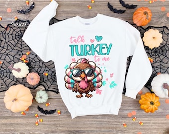 May include: White sweatshirt with a cartoon turkey wearing sunglasses. The text "talk TURKEY to me" is in teal and pink. The design includes a turkey illustration and decorative leaves. The sweatshirt is surrounded by Halloween decorations.