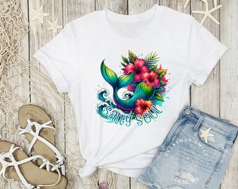 May include: White t-shirt featuring a colorful mermaid tail design with the text "Salty Soul". The mermaid tail is green, blue, and purple, surrounded by pink hibiscus flowers and green leaves. The shirt is paired with denim shorts and sandals.