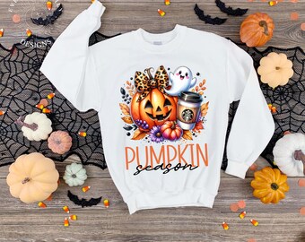 May include: White sweatshirt with a colorful pumpkin-themed graphic. The design includes a jack-o'-lantern, ghost, coffee cup, and the words "PUMPKIN season" in orange. The sweatshirt is surrounded by pumpkins and Halloween decorations.