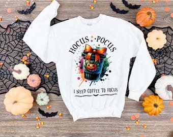 May include: White sweatshirt with a Halloween-themed graphic. The design features a coffee cup with a pumpkin, a bow, and the words "Hocus Pocus" and "I need coffee to focus". Decorative pumpkins and bats surround the sweatshirt.