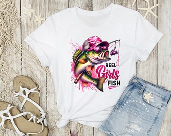 May include: White t-shirt featuring a colorful graphic of a fish wearing sunglasses and a hat, holding a fishing rod. The text "Reel Girls Fish" is printed in pink. The shirt is paired with denim shorts and sandals.