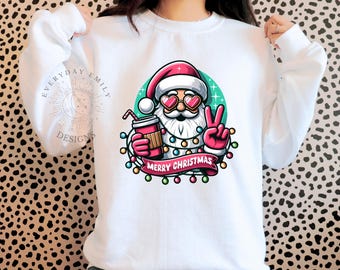 May include: White sweatshirt featuring a cartoon Santa Claus with heart-shaped sunglasses, holding a drink and giving a peace sign. The design includes a banner that reads "MERRY CHRISTMAS" and is surrounded by colorful lights.