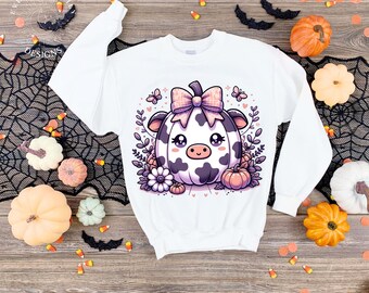 May include: White sweatshirt featuring a cartoon cow-pumpkin design with a pink bow, purple spots, and floral accents. The design is surrounded by leaves, flowers, and small pumpkins, perfect for Halloween or fall. The sweatshirt is displayed on a wooden surface.