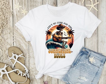 May include: White t-shirt featuring a skeleton playing a guitar on a wave, with a sunset backdrop. Text reads "Ain't no cure for the Summertime Blues." The design includes palm trees and a cowboy hat. A casual summer tee.