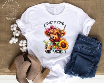 May include: White t-shirt with a colorful graphic of a Highland cow drinking iced coffee, a sunflower, and the text "Fueled by Coffee and Anxiety." The shirt is paired with blue jeans and tan ankle boots.