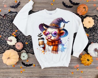 May include: White sweatshirt featuring a whimsical illustration of a cow wearing a witch hat, glasses, and a scarf, holding a coffee cup. The design includes bats and the text "EVERYDAY EM DESIGNS". Halloween-themed apparel.