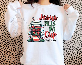 May include: White sweatshirt with a graphic of a coffee cup decorated with a red bow and Christmas lights. The text "Jesus Fills My Cup" is in red and green, with "Psalm 23:5" below. The design is on the front of the sweatshirt.