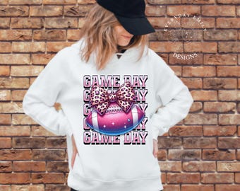 May include: White hoodie with a football graphic and the text "GAME DAY". The football is pink and blue with a leopard print bow. The hoodie has long sleeves and a hood.