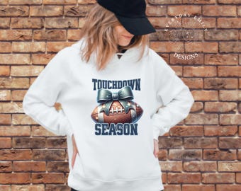 May include: White hoodie with a football graphic and the words "Touchdown Season". The football is brown with a blue bow and silver accents. The hoodie is worn by a person wearing a black baseball cap.
