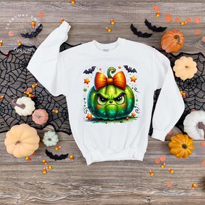Puede incluir: Sudadera blanca con un diseño de calabaza verde de dibujos animados con una cara enfadada, un lazo naranja y motivos de murciélagos y estrellas. La parte superior de manga larga está rodeada de decoraciones de Halloween.