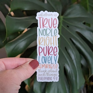 May include: A colorful sticker with the words "whatever is TRUE NOBLE RIGHT PURE LOVELY ADMIRABLE think about such things PHILIPPIANS 4:8" in a vertical layout. The sticker is held by a hand with a maroon nail polish.