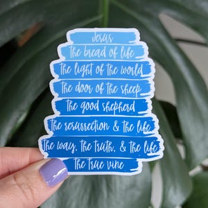 May include: A blue and white sticker with the words "Jesus" and other phrases like "The bread of life" and "The light of the world". The sticker is held by a hand with a purple nail polish.