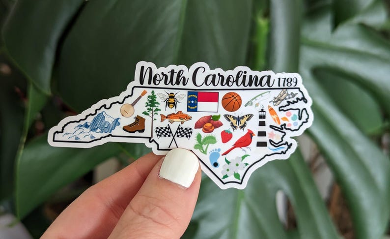 May include: A white sticker shaped like the state of North Carolina with the text "North Carolina 1789" and various icons representing the state, including a bee, a fish, a basketball, a lighthouse, a cardinal, and a mountain range.