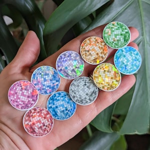 May include: A collection of nine iridescent disco ball stickers in various colors, including green, blue, silver, gold, and pink. The stickers are arranged in a semi-circle on a hand.