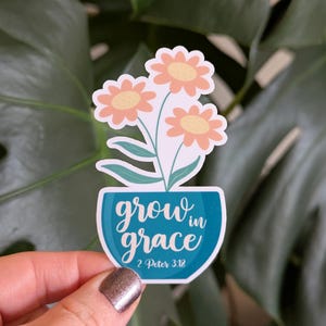 May include: A sticker featuring a floral design with three peach-colored flowers and green leaves in a teal vase. The text "grow in grace" is written in white script, with "2 Peter 3:18" below.