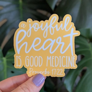 May include: A yellow sticker with the text "a joyful heart is good medicine Proverbs 17:22" in white script. The sticker is held in front of a background of green leaves.