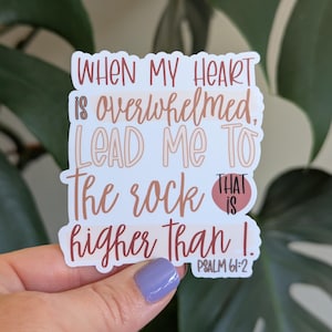 May include: A sticker with the text "When my heart is overwhelmed, lead me to the rock that is higher than I. Psalm 61:2" in a mix of brown and white fonts. The sticker is held by a hand with a purple nail polish.