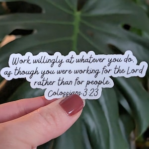 May include: A white sticker with black text that reads "Work willingly at whatever you do, as though you were working for the Lord rather than for people. Colossians 3:23." The sticker is held in front of a green leafy background.