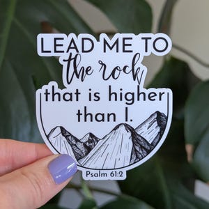 May include: A white sticker with the text "LEAD ME TO the rock that is higher than I." and "Psalm 61:2." The sticker features a black and white illustration of mountains. The sticker is held in front of a green leafy background.