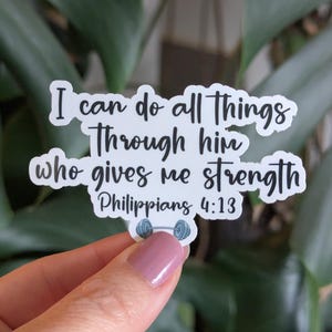 May include: A white die-cut sticker featuring the black text I can do all things Through him who gives me strength Philippians 4:13 in a cursive script font, with a small illustration of a blue dumbbell centered at the bottom edge.