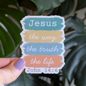 May include: A sticker with the words "Jesus the way the truth the life John 14:6" in white text on colorful horizontal bars. The bars are in shades of blue, yellow, and orange, with a white border. The sticker is held by a hand.