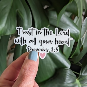 May include: A white sticker with the text "Trust in the Lord with all your heart Proverbs 3:5" in black script. A pink heart is below the text. The sticker is held in front of green leaves.