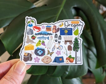 Puede incluir: Una pegatina blanca con forma del estado de Oregón con varias ilustraciones de cosas asociadas al estado, incluyendo una foca, un árbol, un pez, una montaña, un pájaro, una tienda de campaña, un carro, un hongo, una avellana y el texto "Oregon" y "1859".