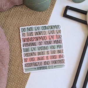 May include: A rectangular sticker with the text "DO NOT BE CONFORMED TO THIS WORLD, BUT BE TRANSFORMED BY THE RENEWAL OF YOUR MIND...ROMANS 12:2". The sticker has pastel color stripes and is on a neutral background.