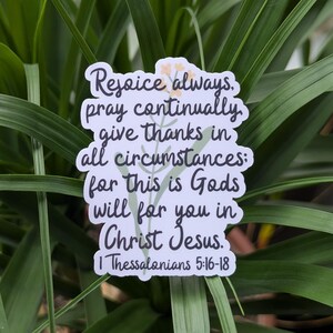 May include: A white sticker with the text "Rejoice always, pray continually, give thanks in all circumstances: for this is Gods will for you in Christ Jesus. 1 Thessalonians 5:16-18" in black script. The sticker has a floral design and is set against a background of green leaves.