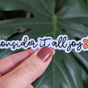 May include: A white sticker with the words "Consider it all joy" in black script, with a small orange flower graphic. The sticker is held in front of a green leafy background.