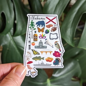 May include: A white sticker shaped like the state of Alabama with various illustrations of things associated with the state, including a peach, a rocket, a butterfly, a football, a bear, a pecan, a red flower, a bird, a bridge, a catfish, a boat, a turkey, a mask, a capital building, and the state flag. The text "Alabama" and "1819" are also included on the sticker.