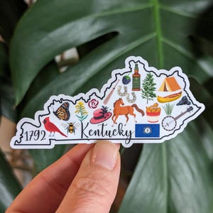 May include: A white sticker shaped like the state of Kentucky with various icons representing the state, including a cardinal, a horse, a banjo, a bottle of bourbon, a derby hat, and the text "Kentucky".