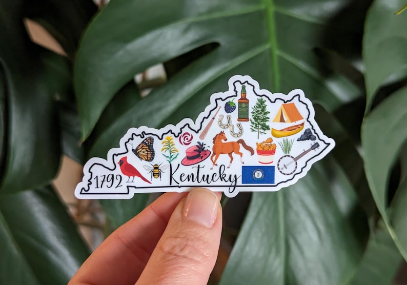 May include: A white sticker shaped like the state of Kentucky with various icons representing the state, including a cardinal, a horse, a banjo, a bottle of bourbon, a derby hat, and the text "Kentucky".