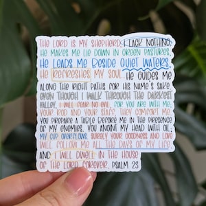 May include: A white sticker with colorful text from Psalm 23. The text includes phrases like "The Lord is my shepherd" and "I will dwell in the house of the Lord forever." The sticker is held in front of a green leafy background.