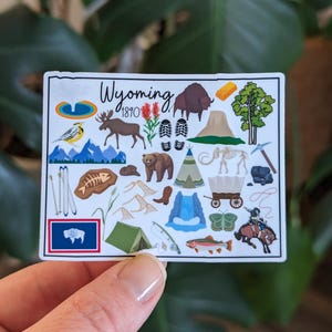 May include: A white sticker with a colorful illustration of various symbols representing Wyoming, including a bison, a mountain range, a tipi, a cowboy, a fish, a fossil, a gold nugget, and the text "Wyoming 1810".