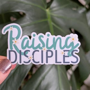 May include: A white sticker with the text "Raising Disciples" in teal lettering with white flowers.