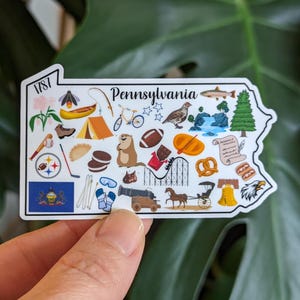 May include: A white sticker in the shape of Pennsylvania, featuring illustrations of state symbols and landmarks. Includes a canoe, a bicycle, a football, a pretzel, and the state flag. The word "Pennsylvania" is written in script.