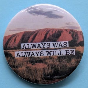 May include: A round button featuring a photograph of a large red rock formation with a cloudy sky in the background. The text "ALWAYS WAS ALWAYS WILL BE" is printed in black on a white background.